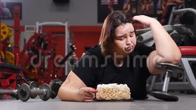 年轻肥胖妇女躺在健身房地板上，吃着美味的高热量蛋糕。 缺乏意志力。 不健康食品